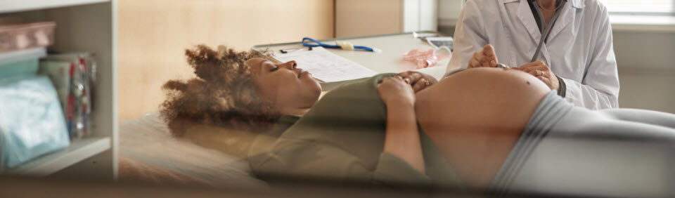 gynecologist with stethoscope checking fetal heart tones while pregnant Black female patient